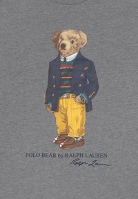 T-shirt grigia con un orso in un blazer blu, maglione a righe, pantaloni a righe gialle e scarpe marroni, con "POLO BEAR by RALPH LAUREN" sottostante.