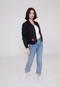 Sort bomberjakke med ribbede manchetter og kant, lynlåslukning og orange labeldetalje, parret med lyseblå denimjeans og hvide sneakers.