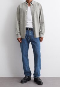 Ein Mann trägt ein hellgraues Hemd über einem weißen T-Shirt, blaue Jeans mit schwarzem Gürtel und schwarze Mokassins und steht vor einer weißen Wand.