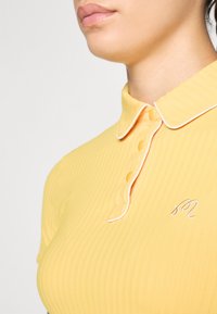 Polo shirt giallo a costine con patta abbottonata e rifinitura del colletto bianca, con una piccola lettera "M" ricamata sul petto.
