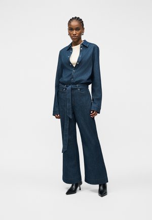 Woman wearing a dark blue denim shirt and matching wide-leg pants with a belt, white top underneath, and black heeled ankle boots.