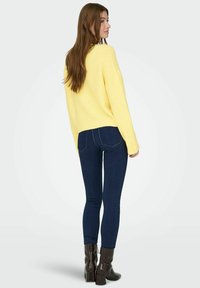 Pull jaune en maille à coupe décontractée, associé à un jean skinny bleu foncé et des bottines marron texturées. Le modèle est présenté de dos.