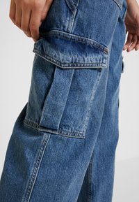 Pantalones cargo de mezclilla azul con múltiples bolsillos laterales, costuras visibles y un ajuste holgado, con una textura ligeramente desgastada cerca de las costuras.