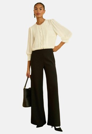 Helle cremefarbene Bluse mit Puffärmeln und gewelltem Detail, kombiniert mit hoch taillierten schwarzen, weit geschnittenen Hosen und einer schwarzen Handtasche.