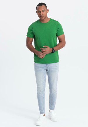 Camiseta de algodón verde con cuello redondo, mangas cortas, combinada con jeans ajustados de lavado claro y zapatillas blancas, con detalles mínimos y textura suave.
