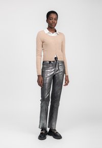 Beige Strickcardigan mit Knöpfen und einer taillierten Passform, kombiniert mit metallisch silbernen Schlagjeans. Schwarze Slipper mit Kettendetails runden das Outfit ab.