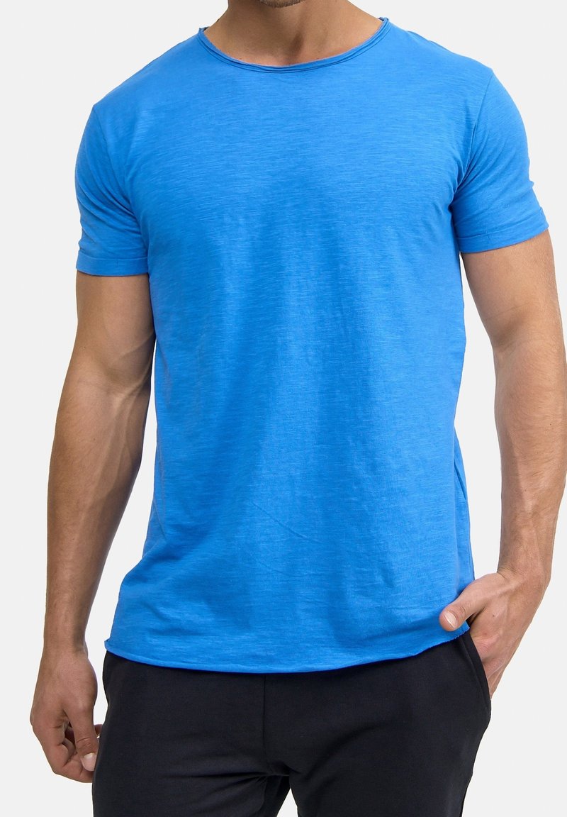 Homme portant un t-shirt à manches courtes bleu vif uni et un pantalon noir, une main dans la poche, sur fond blanc.