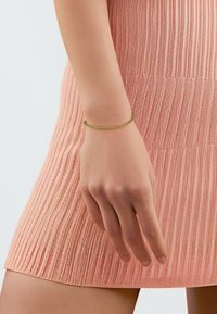 Goudkleurige vlechtarmband om een hand, gedragen met een gebreide roze jurk met een gestructureerd patroon en verticale ribbels.