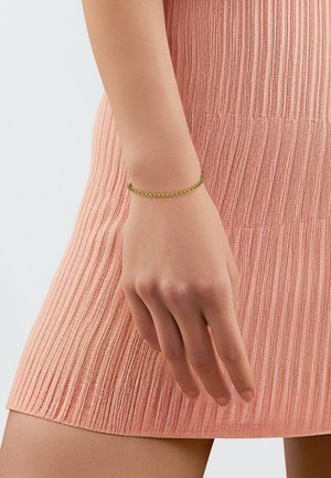 Goudkleurige vlechtarmband om een hand, gedragen met een gebreide roze jurk met een gestructureerd patroon en verticale ribbels.