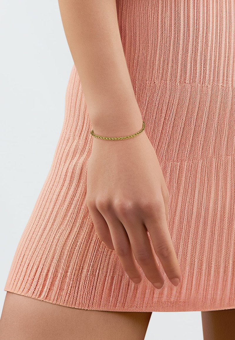 Goudkleurige vlechtarmband om een hand, gedragen met een gebreide roze jurk met een gestructureerd patroon en verticale ribbels.