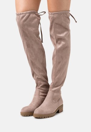 Bottes hautes en daim couleur taupe clair, avec bouts ronds, talons bas carrés, semelles crantées et lacets ajustables en haut, portées sur des jambes nues.