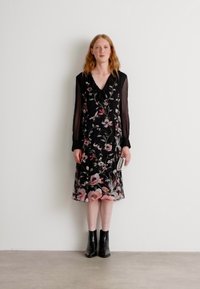 Schwarzes Blumenkleid mit pinken und grünen Mustern, einem V-Ausschnitt und transparenten Ärmeln. Getragen mit schwarzen Knöchelboots.