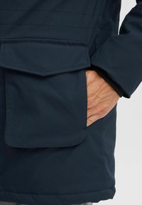 Marineblauer wasserdichter Anorak mit einer großen Klappentasche, glatter Stoffoberfläche und verstellbarem Ärmelbündchen. Hand ruht in der Tasche.