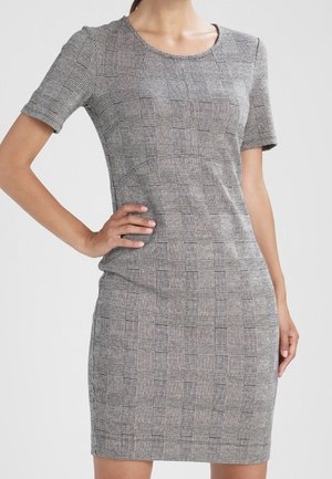 Frau trägt ein figurbetontes, kurzärmeliges, knielanges graues Kleid mit einem schwarz-weiß karierten Muster, steht mit einer Hand in der Hüfte.