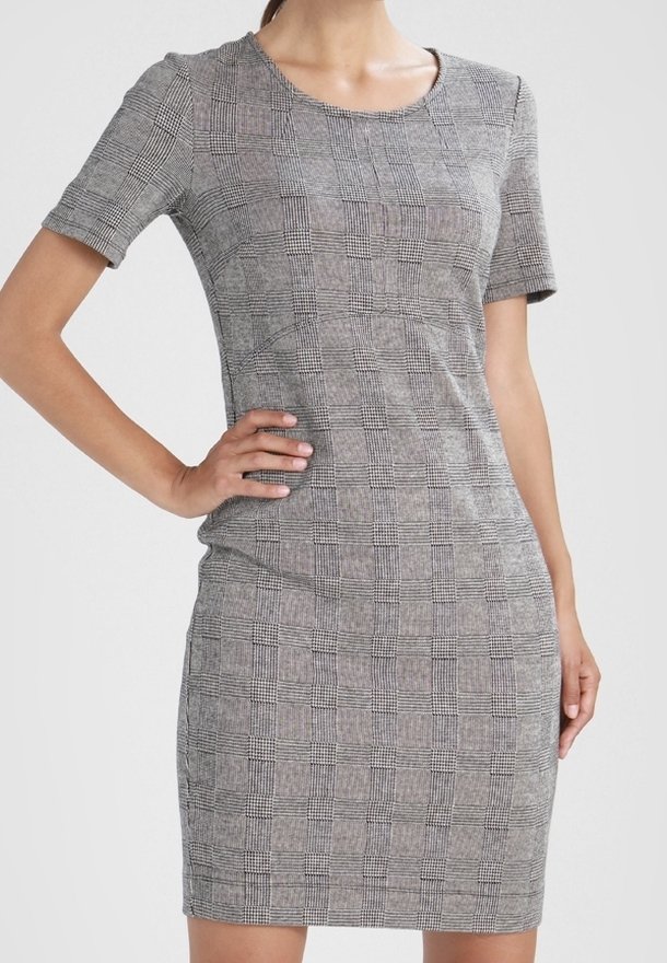 Femme portant une robe grise ajustée, à manches courtes, longueur genou, avec un motif à carreaux noir et blanc, debout avec une main sur la hanche.