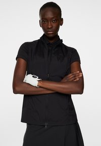 Svart ärmlös zip-up jacka med ståkrage, tillverkad av lättviktig tyg, i kombination med en vit golfhandske på modellens korsade armar.