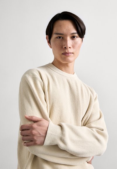 Pull beige à côtes avec un col ras du cou et des manches longues, présentant un tissu texturé. La personne a les bras croisés, obscurcissant partiellement les détails.