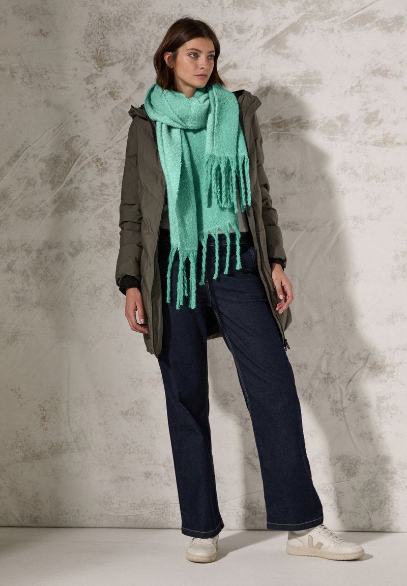 Parka verde oliva voluminoso su un top grigio chiaro, abbinato a jeans a gamba larga di colore blu scuro e sneakers bianche. Una sciarpa di un verde menta con frange adorna il collo.