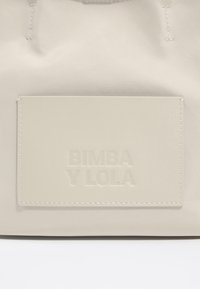 Borsa beige con una texture liscia, caratterizzata da una patch logo testurizzata e dettagli di cucitura. Design semplice e funzionale, privo di abbellimenti.