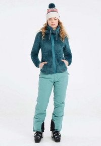 Mėlynas pūkų džemperis su tekstūruotu audiniu ir užtrauktuku, derinamas su šviesiai mėlynomis sniego kelnėmis su juodais akcentais ir slidinėjimo batais.