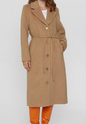 Frau in einem langen, kamelfarbenen, knöpfbaren Mantel mit Gürtel, weißem Oberteil und leuchtend orangefarbenen Stiefeln, die vor einem schlichten Hintergrund steht.