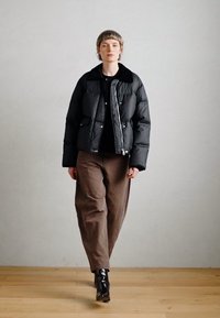 Črna puffer jakna s skrajšano zasnovo, z zadrgo, srebrnimi dodatki in mehak ovratnik. V kombinaciji z rjavimi hlačami in črnimi škornji.