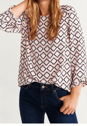 Femme portant un chemisier rose à manches longues avec un motif géométrique noir en forme de losanges et un jean bleu foncé, main dans la poche, posant avec une posture détendue.