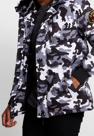 Kurtka moro w czarno-biało-szarym kolorze z wieloma kieszeniami i żółtą naszywką na rękawie, noszona na ciemnej koszulce.