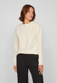 Lichtbeige sweatshirt met een ronde hals, lange mouwen en geribde manchetten, gecombineerd met zwarte broek. Zachte textuur, ontspannen pasvorm.
