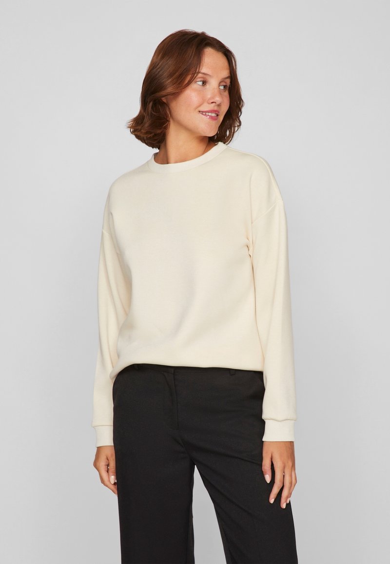 Sudadera de color beige claro con cuello redondo, mangas largas y puños acanalados, combinada con pantalones negros. Textura suave, corte relajado.