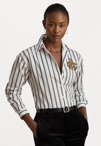 Cămașă albă cu dungi negre, cu un model brodat în aur pe piept, asortată cu pantaloni negri și o curea.