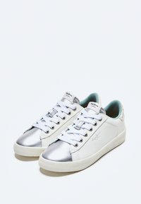 Zapatillas blancas con acentos metálicos plateados, cordones blancos y una suela de goma texturizada. Cuenta con un interior acolchado en azul claro.