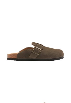 Brun ruskind slip-on træsko med justerbar sidespænde og korksål, set fra den indvendige side på hvid baggrund.