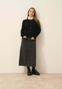 Svart stickad cardigan med knappar, matchad med en mörkgrå denim-maxikjol. Den kompletta looken inkluderar svarta skor och en vit polotröja.