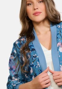 Zils ziedu kimono žakete ar rozā un baltām ziedu rakstiem, viegls audums, atvērts priekšpuse, kontrastējošs gaiši zils apmales izšuvums un zem tā valkāts V-veida kakla tops.