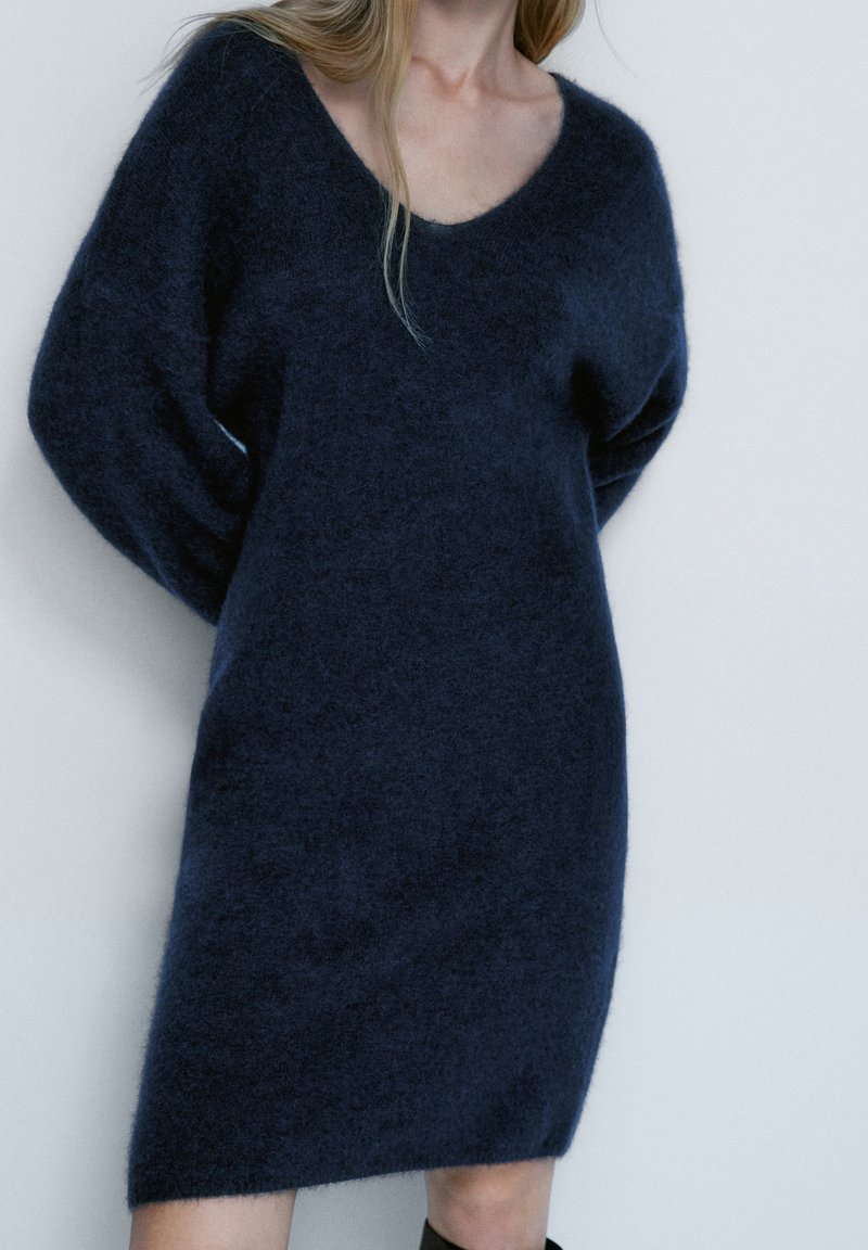 Femme portant une robe en tricot ample et duveteuse de couleur bleu foncé avec des manches longues et un décolleté arrondi, se tenant contre un mur blanc uni.