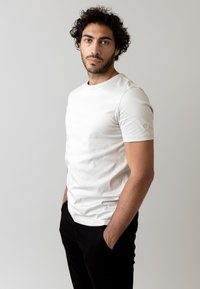 T-shirt bianco in cotone con scollo rotondo e maniche corte, caratterizzato da un logo discreto sulla manica sinistra, abbinato a pantaloni neri.