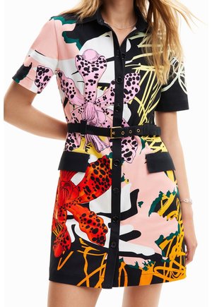 Kobieta w krótkim, zapinanym na guziki sukience z czarnym paskiem i wyrazistym kwiatowym wzorem w kolorach różowym, czerwonym, żółtym i czarnym.