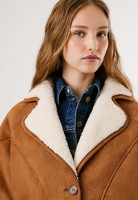 Cappotto in suede marrone con colletto in pelliccia sintetica crema, dotato di chiusura con bottoni e una camicia di jeans sotto. La texture è soffice e liscia.
