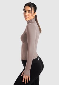 Langærmet top i lys taupe, lavet af strækbart stof. Har en høj krave og sømløs design, parret med sorte leggings.