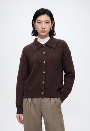 Jeune femme portant un cardigan marron foncé boutonné avec un large col sur un haut blanc et un pantalon plissé beige, debout avec une main dans la poche.