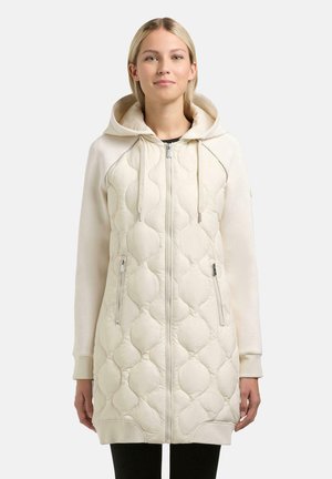 Kobieta ubrana w długą, kremową pikowaną bluzę z kapturem na zamek, z troczkami i przednimi kieszeniami na zamek, stojąca na jednolitym tle.