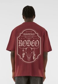 Maroon cotton t-shirt with a graphic design featuring a skull and feathers, displaying the word "RODEO" in bold text at the back.