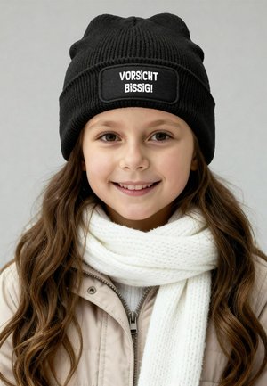 Black knit beanie with a rectangular patch featuring white text. Paired with a white textured scarf and light-colored zip-up jacket.