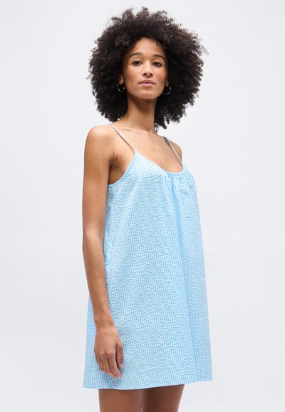 Γυναίκα με σγουρά μαλλιά που φοράει ανοιχτό μπλε μαν sleeveless φόρεμα με λεπτές τιράντες, στέκεται μπροστά σε ένα απλό φωτεινό φόντο.