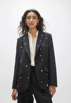 Mujer con blazer negro con tachuelas redondeadas y coloridas, camisa blanca, pantalones negros y pendientes cuadrados de oro sobre un fondo blanco liso.