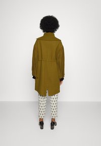 Olivgrön kappa med texturerade fransar på ärmarna och hög krage, matchad med mönstrade vita byxor med geometrisk design. Svarta skor.