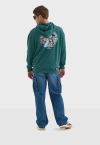Sudadera verde con un diseño gráfico circular en la espalda, combinada con unos jeans cargo azules, exhibiendo un corte relajado y múltiples bolsillos.