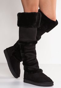 Svarta knähöga stövlar med en mjuk, syntetisk pälsmudd och i mockamaterial. Designen har en slapp textur och en platt gummisula.