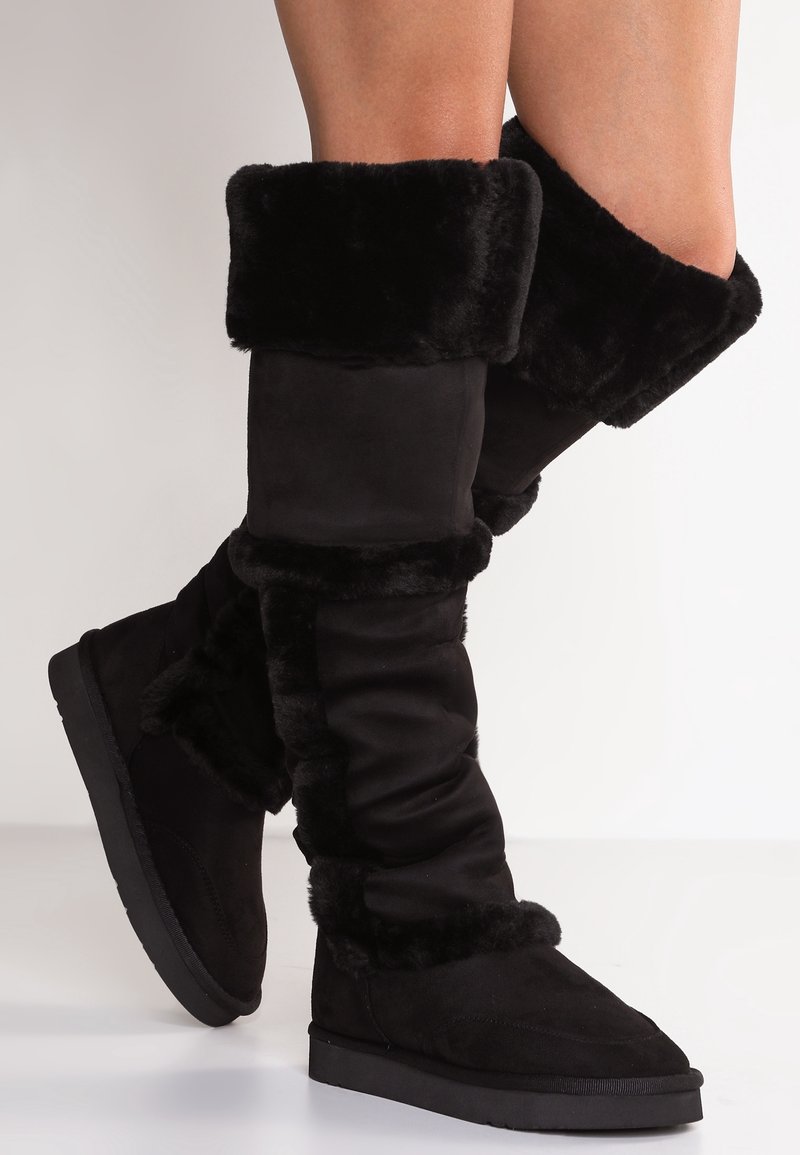 Svarta knähöga stövlar med en mjuk, syntetisk pälsmudd och i mockamaterial. Designen har en slapp textur och en platt gummisula.